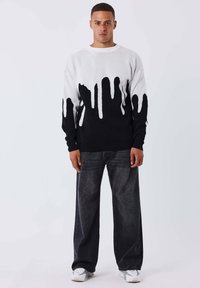 Suéter de punto en blanco y negro con patrones de goteo, con mangas largas negras y un cuello redondo, combinado con pantalones de mezclilla oscuros y zapatillas blancas.