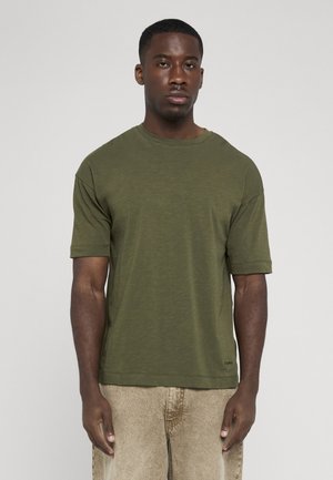 Mand med kort hår iført en olivengrøn kortærmet t-shirt og beige bukser, står mod en ensfarvet lys baggrund.