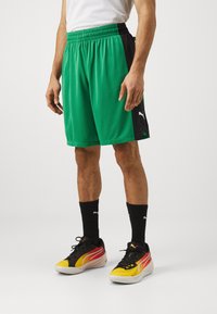 Žalios sportinės šortai su juodomis šoninėmis juostelėmis, elastingu juosmeniu ir Puma logotipu. Dėvimi su juodais ir geltonais bateliais bei juodomis kojinėmis.