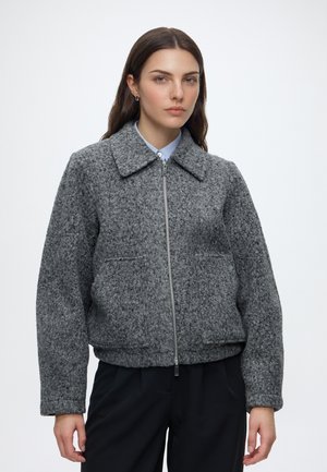 Femme portant une veste zippée texturée grise avec un col, associée à un pantalon noir, debout devant un fond blanc uni.