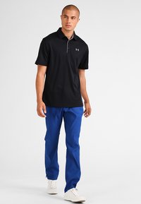 Camisa polo negra con cuello, acentos grises, combinada con pantalones azules y zapatillas blancas, mostrando un estilo atlético casual.
