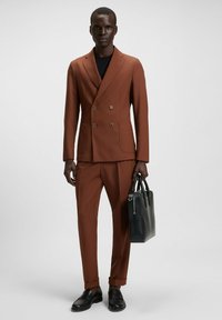 Costume marron à double boutonnage avec deux poches avant, associé à un haut noir. Le modèle tient un sac noir et porte des chaussures noires.