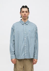 Prúžkovaná košeľa na gombíky v svetlo modrej a bielej farbe, s golierom, dlhými rukávmi a náprsné vrecko. Vyrobená z hladkej, ľahkej tkaniny.