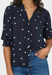 Marineblå skjorte med hvide hjerteprint, med mandarin-krave og elastiske ærmer, parret med blå jeans.