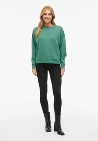 Femme souriante avec de longs cheveux blonds, portant un sweat-shirt vert avec le texte "Minimalisme", un jean skinny noir et des bottes à talons noires, debout.
