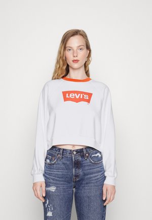 Weißer, cropped Sweatshirt mit rotem Kragen und großem roten Levi's-Logo. Kombiniert mit blauen, zerstörten Jeans im High-Rise-Schnitt.