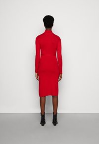 Robe tricot rouge à manches longues avec une silhouette cintrée, un ourlet au genou, un col haut et sans motifs visibles, assortie à des bottines noires.