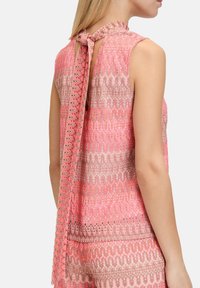 Ärmelloses Oberteil in Rosa und Beige mit Häkelmuster und einer Schleife am Hals. Verfügt über eine strukturierte Oberfläche und einen etwas längeren Rücken.