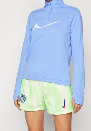 Person iført en lyseblå langærmet top med en hvid swoosh og lysegrønne sportsshorts med et FCB-logo og Nike swoosh.