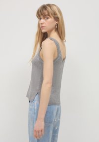 Jeune femme avec de longs cheveux blonds portant un débardeur en tricot argenté et un jean bleu clair, de profil contre un fond uni.