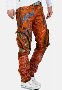 Oranje cargobroek met blauw en roze spetterpatronen, studs, en zijzakken met rits, gecombineerd met witte sneakers.