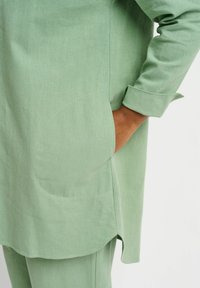 Light green tunic with a relaxed fit, featuring a rounded hem, rolled cuffs, and side pockets. Textured fabric adds visual interest.