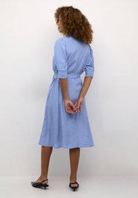 Robe bleu clair, longueur genou, avec de courtes manches bouffantes et une taille ceinturée. Tissu lisse avec une silhouette fluide et un détail au dos.