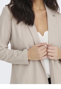 Beige bleiseri, jossa on räätälöity muotoilu, lovilla varustetut läpät, pitkät hihat ja sileä kangas. Yhdistettynä valkoiseen puseroon.