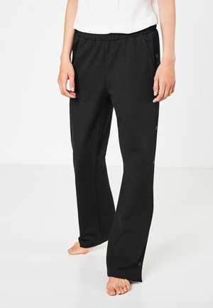 Personne portant un pantalon noir ample avec des boutons sur les côtés et un haut blanc, debout pieds nus sur un sol blanc.