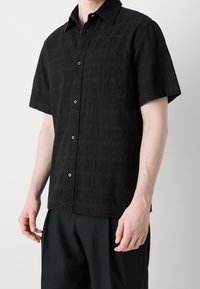 Zwart shirt met korte mouwen, gemaakt van een gestructureerde stof met een geruit patroon, voorzien van een knoopsluiting en een enkele borstzak.