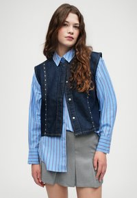 Colete de ganga com detalhes metálicos sobre uma camisa azul às riscas, combinado com uma saia plissada cinza. Inclui botões de pressionar e corte reto.