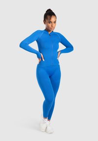 Tætsiddende blå langærmet atletisk top med høj krave og lynlås, parret med matchende blå leggings. Glat, strækbart stof.