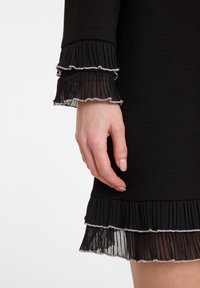 Robe noire avec des poignets en couches plissés et transparents, ornés de bords argentés. Présente une silhouette ajustée et une texture douce. Main partiellement visible.
