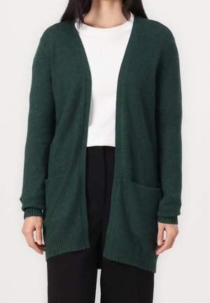 Frau trägt eine dunkelgrüne, offen getragene Strickjacke mit zwei vorderen Taschen über einem weißen Hemd und schwarzen Hosen, steht vor einem einfarbigen Hintergrund.