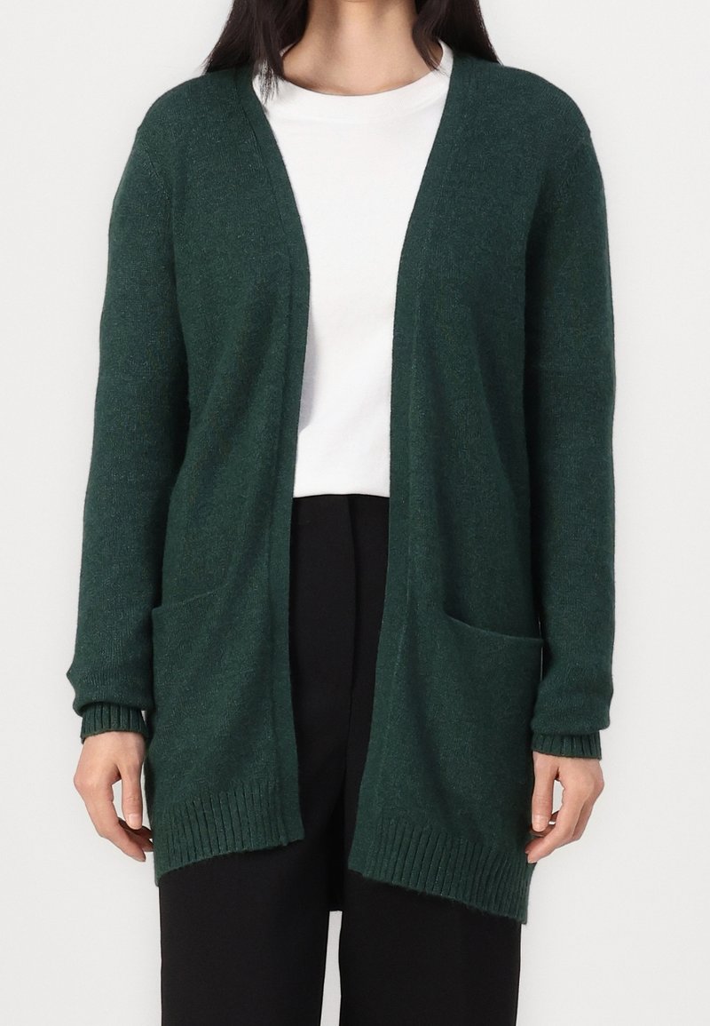 Femme portant un cardigan vert foncé ouvert à l'avant avec deux poches avant, par-dessus une chemise blanche et un pantalon noir, debout devant un fond uni.