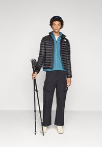 Zwarte pufferjack, blauwe fleece zip-up, zwarte cargo broek en beige schoenen. Houdt trekkingstokken vast, met een gladde, lichte achtergrond.