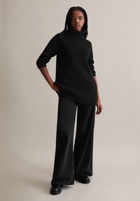 Schwarzer gestrickter Pullover mit hohem Stehkragen, kombiniert mit lockeren schwarzen Denim-Hosen und klobigen schwarzen Plateauschuhen.