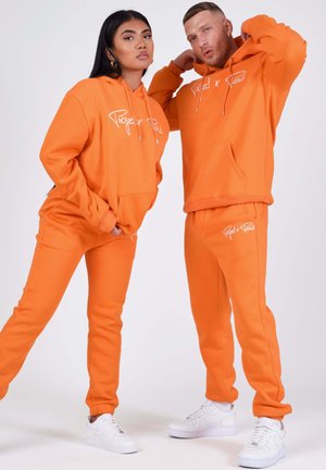 Femme et homme portant des sweats à capuche orange vif assortis et des pantalons de survêtement avec des baskets blanches, posant devant un fond uni.