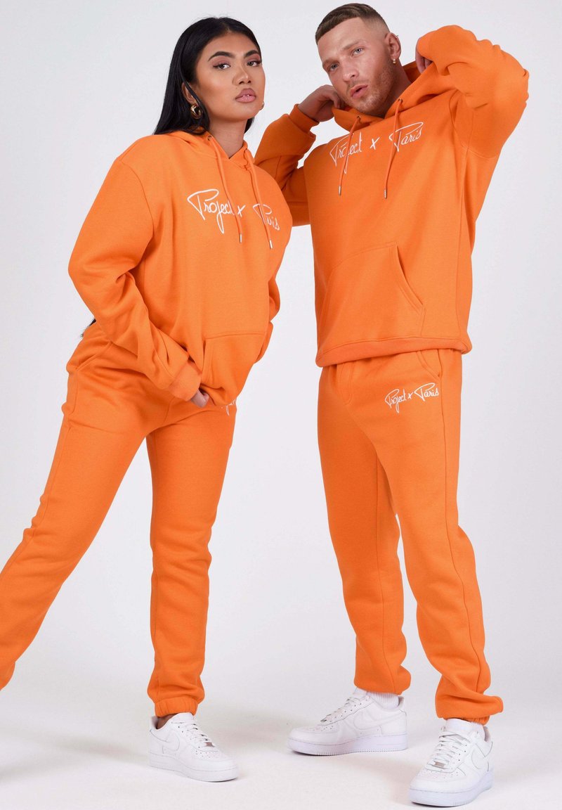 Donna e uomo che indossano felpe con cappuccio e pantaloni della tuta arancione brillante coordinati con scarpe da ginnastica bianche, posano su uno sfondo semplice.