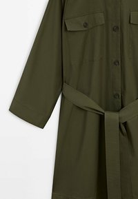 Robe-chemise à manches longues couleur vert olive avec boutons sur le devant, poches poitrine à rabat, et ceinture en tissu assorti nouée à la taille.