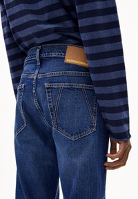 Donkerblauwe jeans met een iets vervaagde afwerking, voorzien van een bruin leren label met de tekst "ARMEDANGELS" en twee achterzakken.