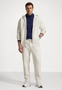 Sweatshirt cinza claro com fecho de correr e cordão, sobreposto a uma camisola de gola alta azul marinho. Calças de fato de treino cinza claro a condizer e sapatilhas brancas desportivas.