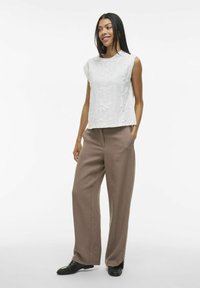 Top blanco sin mangas con patrón floral texturizado, combinado con pantalones beige sueltos. La modelo está de pie con una mano en el bolsillo, usando zapatos negros.