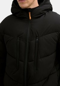 Musta toppatakki hupulla, jossa on vinottain tikkivuori, kaksi vetoketjullista rintataskua ja oranssinvärinen vetoketjun vetolaite.