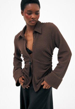 Kobieta w brązowej, teksturowanej koszuli zapinanej na guziki, na czarnej bluzce, z długim czarnym naszyjnikiem z frędzlami i czarną spódnicą, pozująca z rękami przy biodrach.