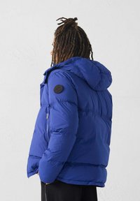 Blauwe pufferjas met een capuchon, gemaakt van een glanzende stof. Bevat ronde patches, ritszakken en een gevoerde textuur.