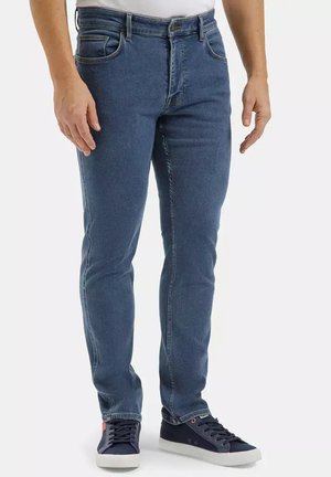 Mężczyzna ubrany w średnio niebieskie obcisłe jeansy i ciemnoniebieskie trampki z białymi podeszwami stoi na jednolitym tle.