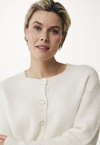 Vit stickad kofta med en strukturerad mönster, rund halsringning och tre stora pärlknappar framtill. Mjukt material.