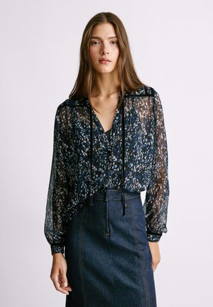 Blusa trasparente a maniche lunghe con motivo floreale nei toni del blu e del bianco, con scollatura a V e dettaglio con cordoncino. Abbinata a una gonna in denim scuro.