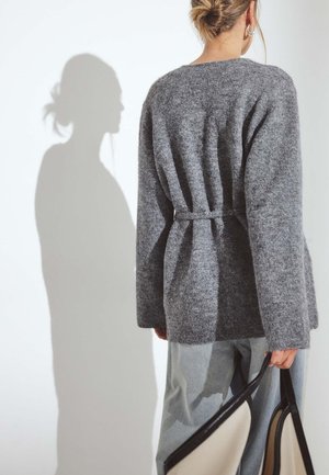 Cardigan grigio in lana con vestibilità rilassata, design avvolgente e maniche lunghe. Notevole lavorazione a maglia testurizzata e un'ombra proiettata sulla parete.