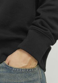 Sudadera negra con puños y bajo acanalados, combinada con vaqueros de denim azules desgastados. Mano reposando en el bolsillo de los vaqueros, mostrando la textura de la tela.