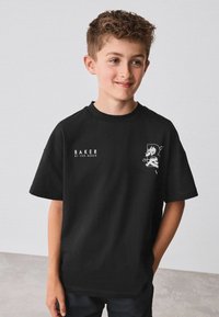 Černé tričko s krátkým rukávem a kulatým výstřihem, s bílým textem "BAKER" a grafickým designem na levé straně. Měkká textilie.