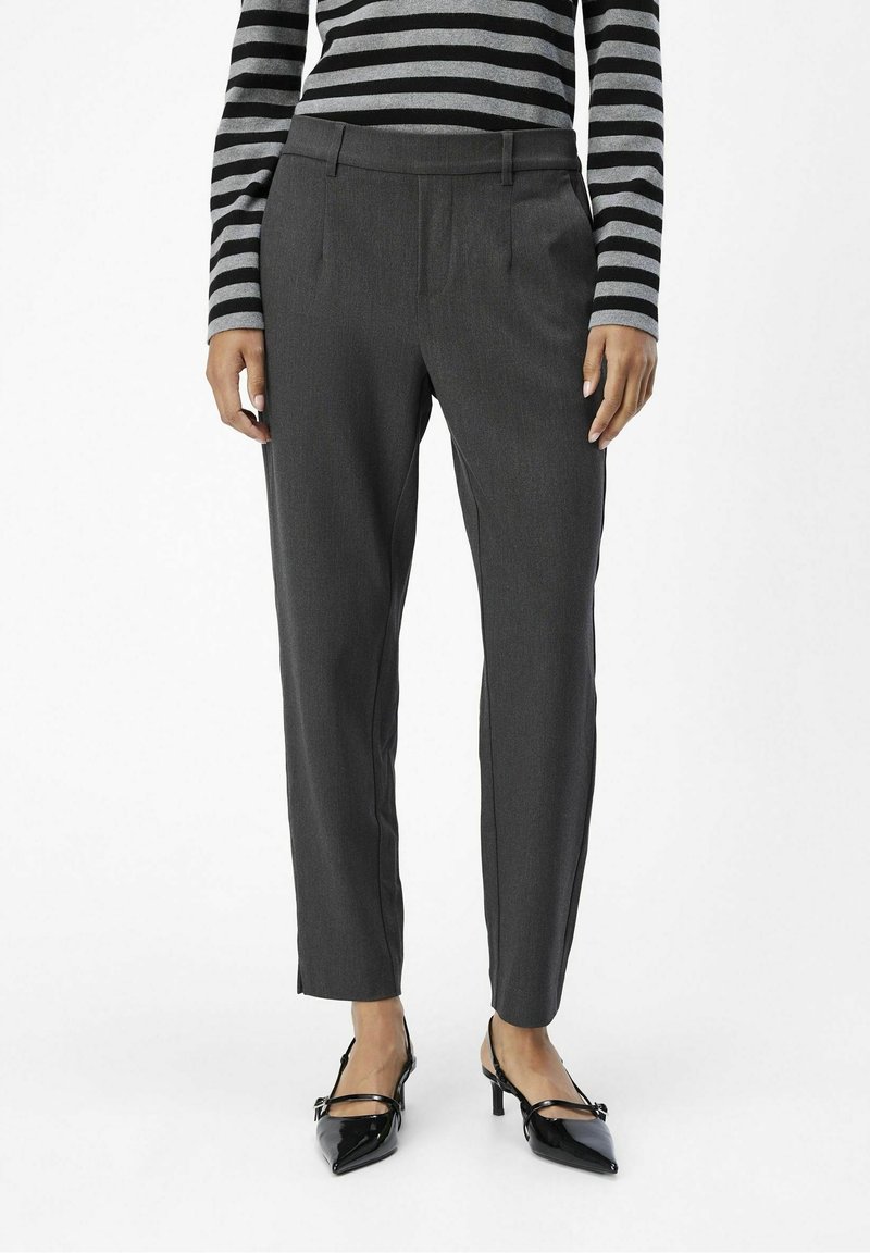 Personne porte un pantalon gris foncé sur mesure, un haut à rayures noires et grises, et des escarpins noirs pointus, debout devant un fond blanc uni.