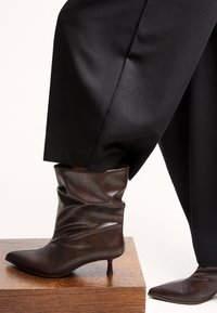 Ghete până la gleznă cu imprimeu de șarpe maro, cu un design lejer, vârf ascuțit și tocul joasă tip stiletto, expuse pe o suprafață de lemn.