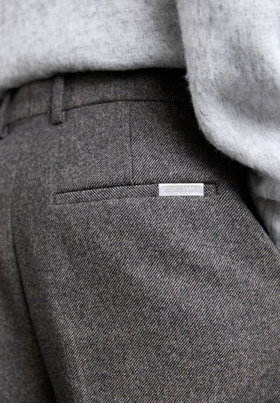 Grå teksturerede stofbukser med en baglomme, der har en sølvmærkat. Ved siden af er en lys grå sweater, delvist synlig ovenfor.