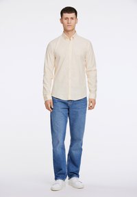Camicia button-up a maniche lunghe di colore giallo chiaro, realizzata in tessuto, abbinata a jeans blu e sneakers bianche; design semplice, taglio aderente, senza fantasie.