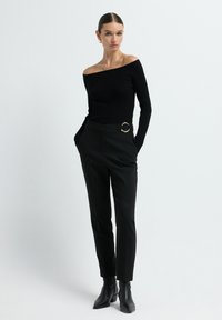 Vrouw in een zwarte off-shoulder ribbeltrui, zwarte op maat gemaakte broek met een gouden gesp riem en zwarte enkelboots, staande met haar handen in haar zakken.