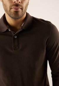 Temno rjava polo majica z dolgimi rokavi iz teksturirane tkanine, z zapenjanjem na gumbe in subtilnim všitim detajlom na ovratniku.