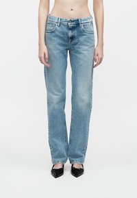 Persoană îmbrăcată cu blugi drepti de culoare albastru deschis și pantofi negri cu vârf ascuțit, stând în fața unui fundal alb simplu.