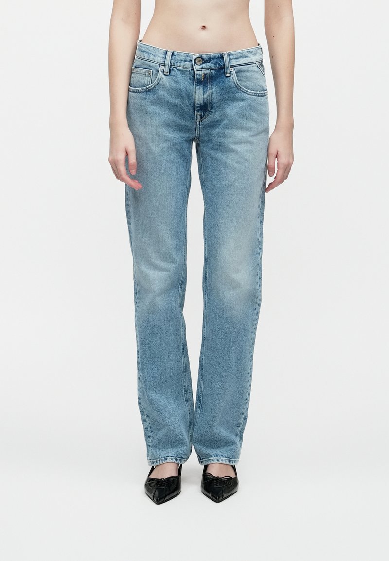 Persoană îmbrăcată cu blugi drepti de culoare albastru deschis și pantofi negri cu vârf ascuțit, stând în fața unui fundal alb simplu.